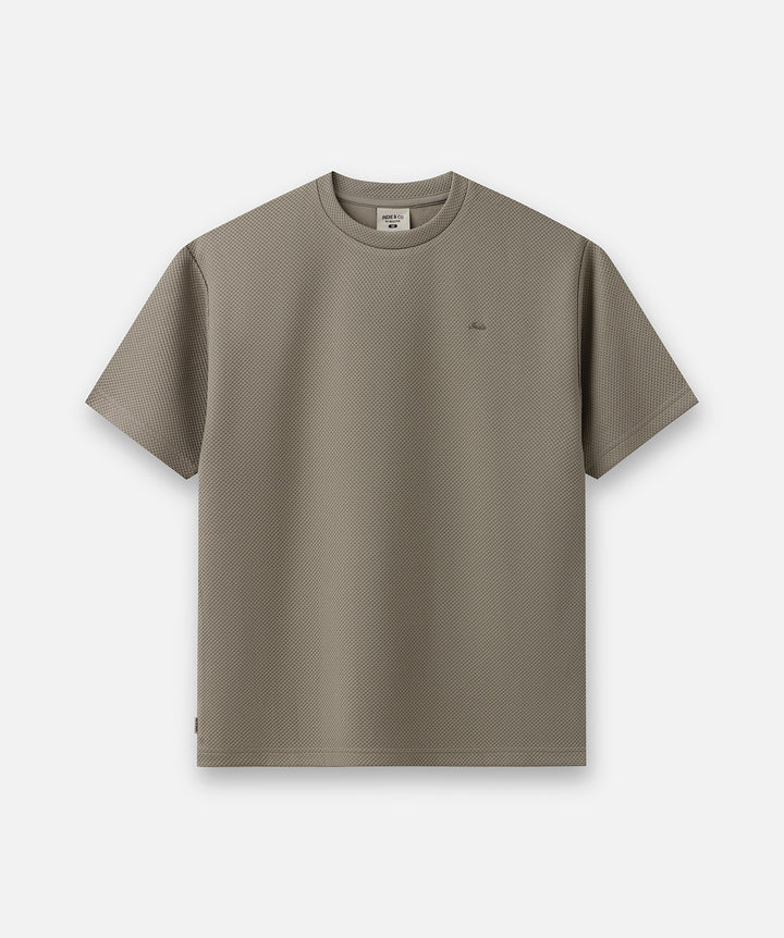 A short-sleeve crewneck t-shirt in a muted taupe-brown color, featuring a subtle textured knit pattern and a small tonal embroidered logo on the left chest, displayed against a clean off-white background.